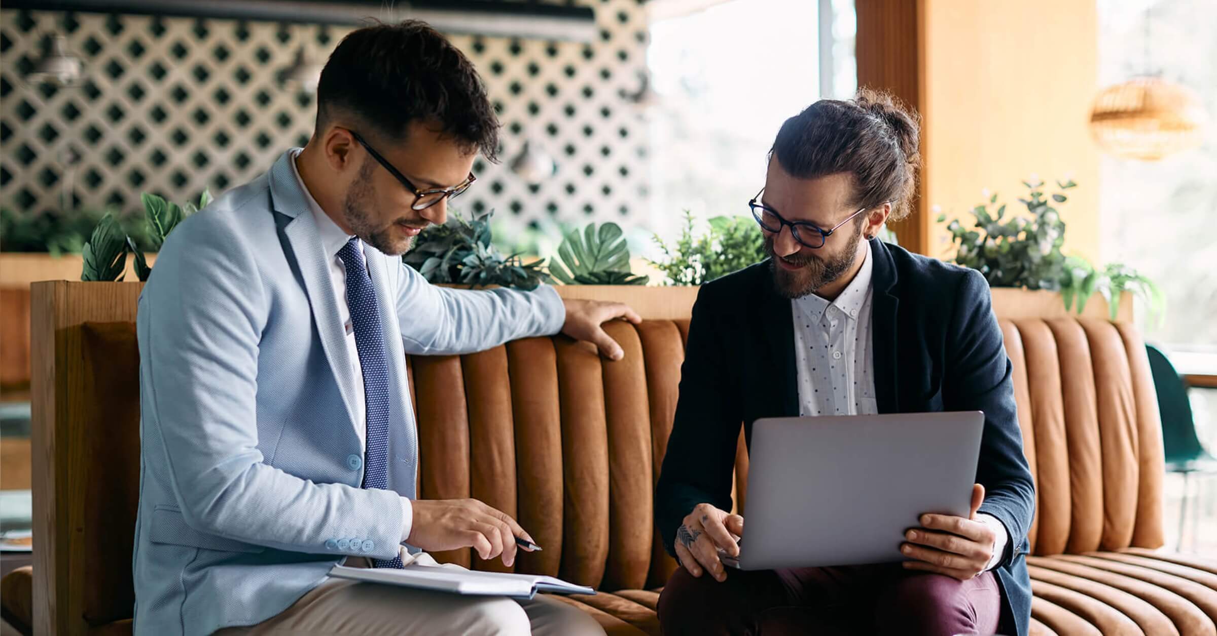 To unge menn med pc og papirer sitter i sofa og samarbeider.