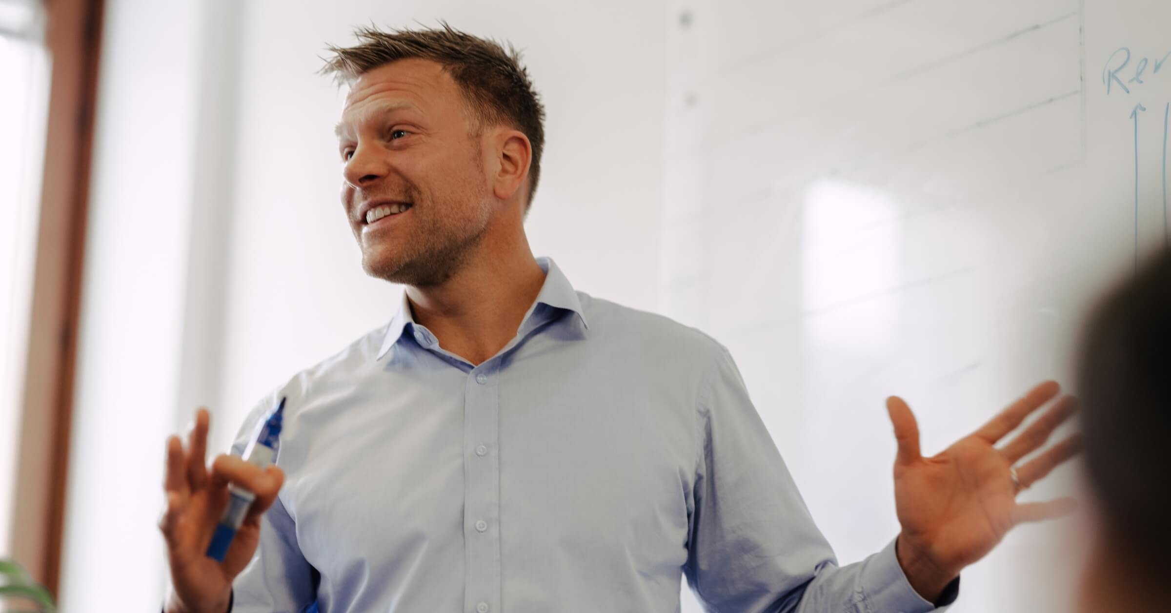 Mann med lys blå skjorte står smilende foran et whiteboard med blå tusj i hånda